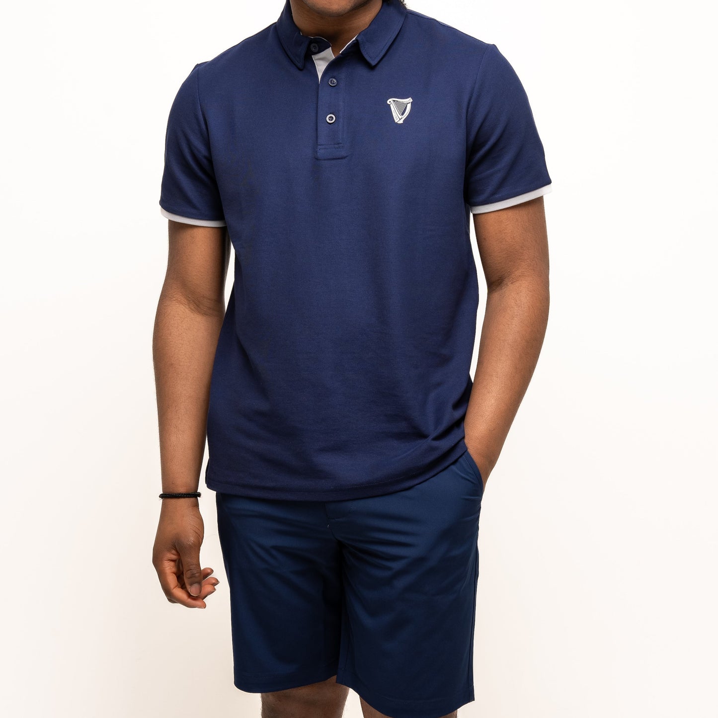 A person in a navy blue polo, Guinness Webstore US Guinness Performance Shorts - Navy with moisture-wicking fabric, and a light gray cap stands with one hand in their pocket against a white background, highlighting an athletic style.