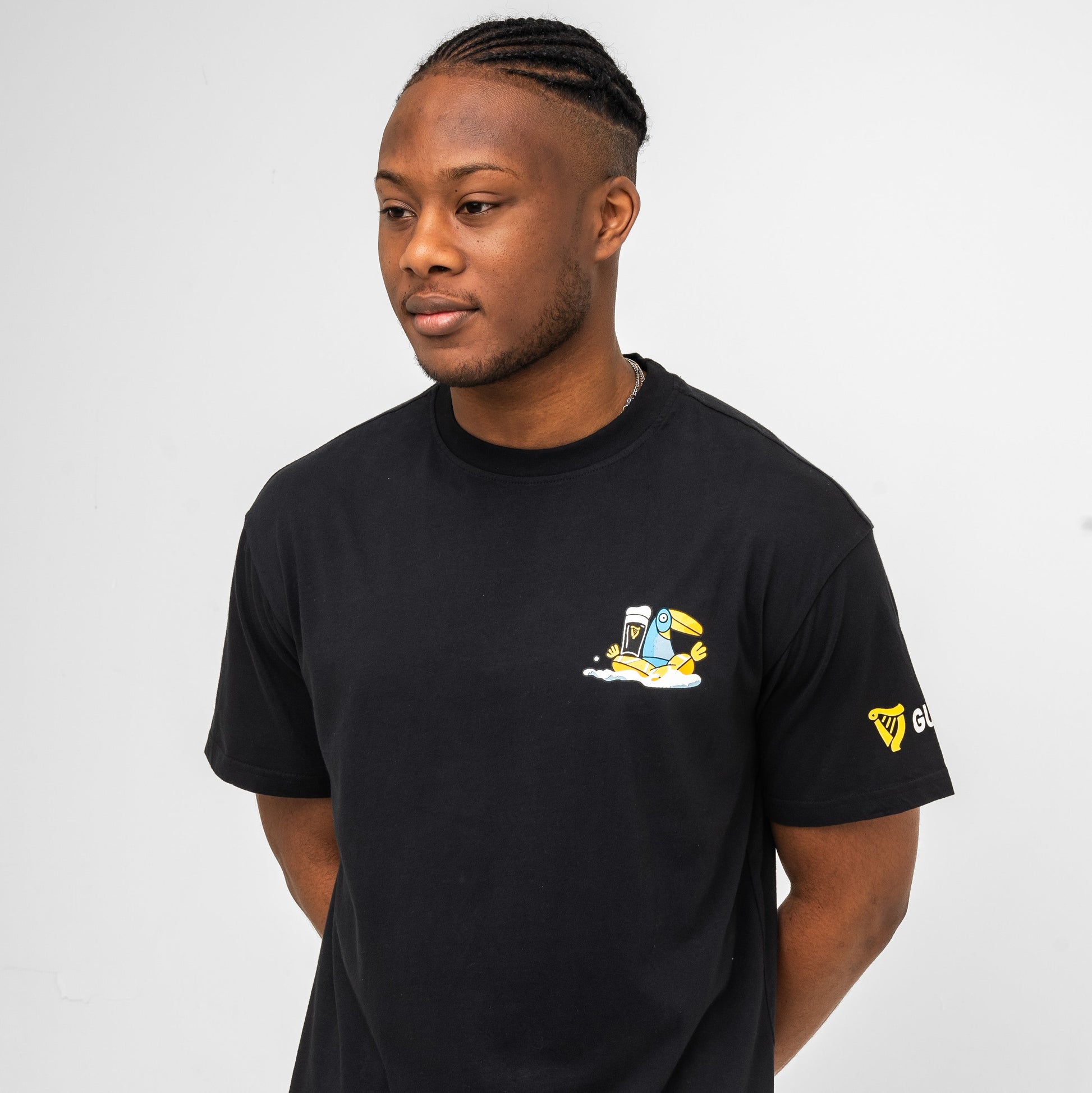 A man with short braids wears the Guinness Seb Curi Oversize Tee - Black by Guinness Webstore US, featuring a small cartoon chest graphic from the Sebastian Curi collaboration, against a plain white background.