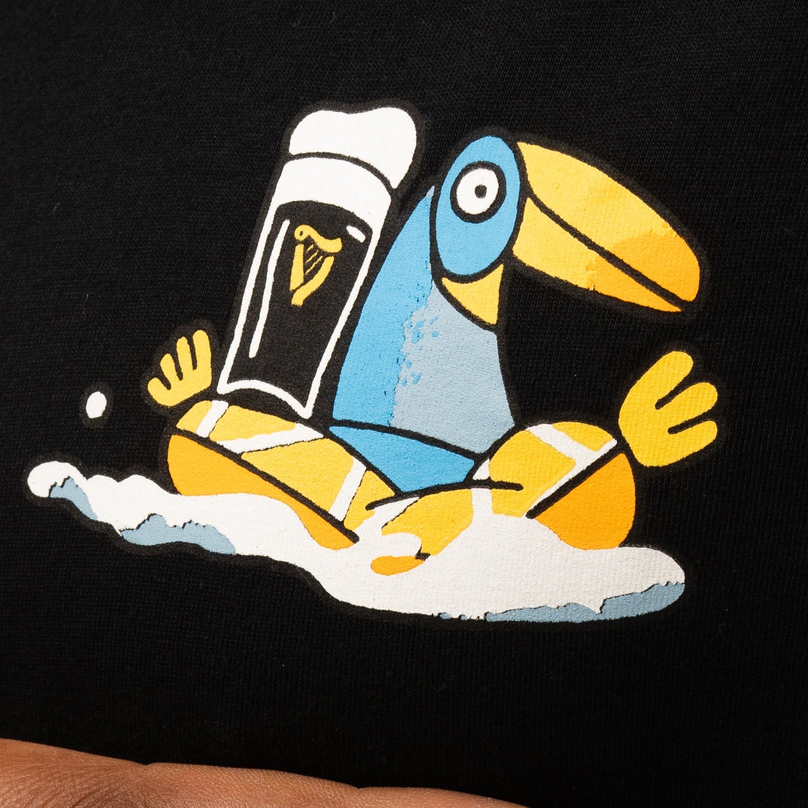 Illustration of a blue and yellow toucan with a pint of Guinness, inspired by Sebastian Curi, on the black Guinness Seb Curi Oversize Tee from Guinness Webstore US.