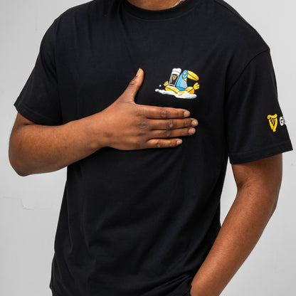 A person in the black Guinness Seb Curi Oversise Tee from Guinness Webstore US, with a small cartoon pint on the chest and a sleeve logo, places their right hand on their chest.