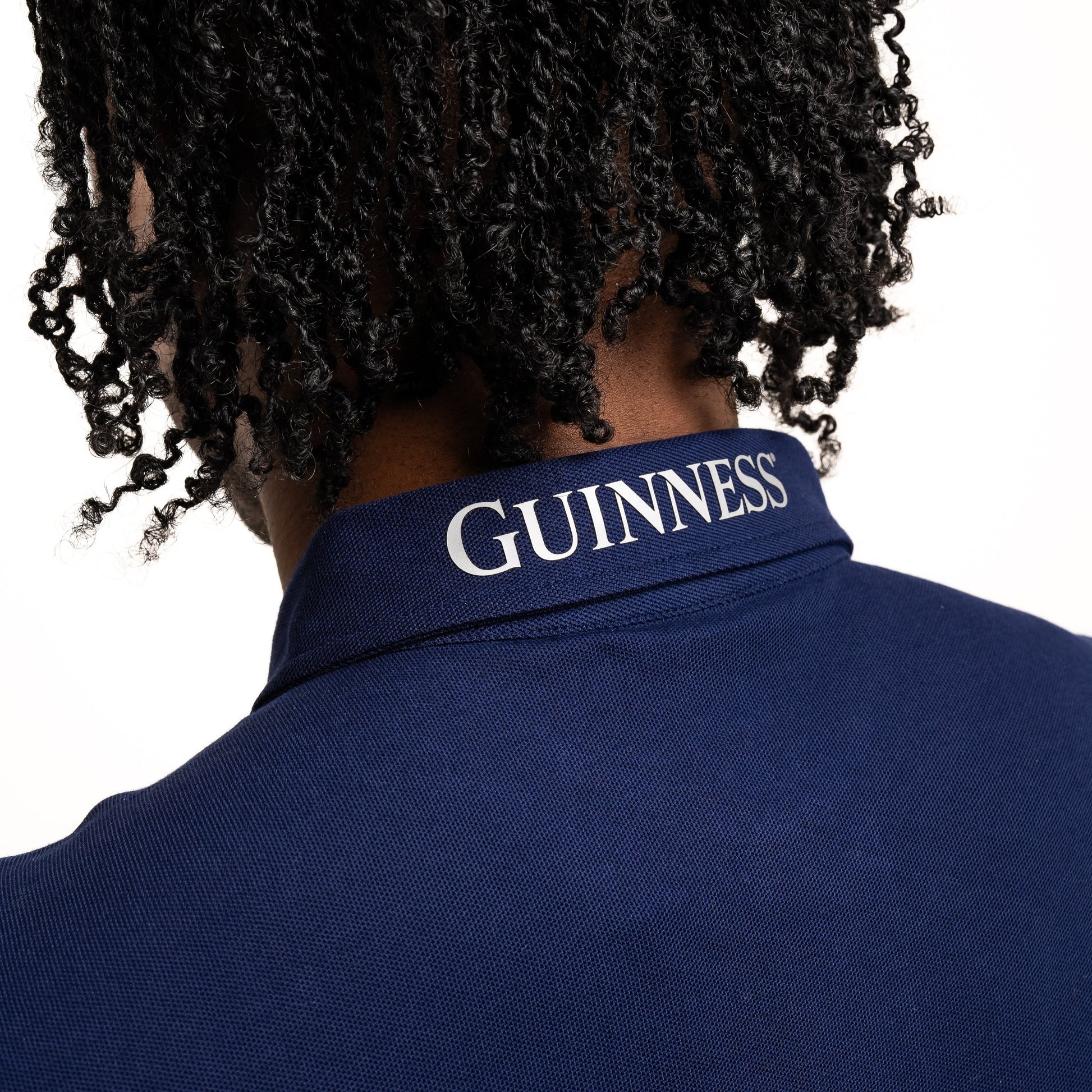 Close-up of a person with short dreadlocks wearing the Guinness Performance Polo - Navy from Guinness Webstore US, featuring "GUINNESS" printed on the collar.
