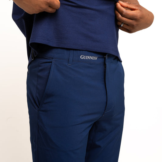A person wearing a blue polo and pants with a "Guinness" logo waistband, lifts their shirt to show Guinness Performance Shorts - Navy by Guinness Webstore US, crafted from moisture-wicking fabric.