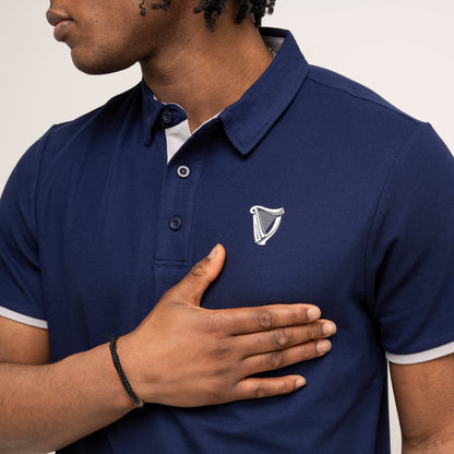 Wearing the Guinness Performance Polo - Navy from Guinness Webstore US, a person places their right hand over their chest, highlighting the polo's classic style and breathable fabric with a white harp emblem.
