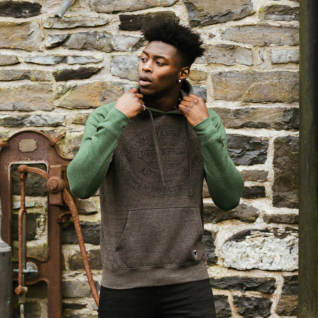 A man wearing a Guinness Grey and Green Hoodie standing in front of a stone wall.