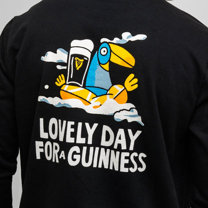 The Guinness Seb Curi Lovely Day Sweatshirt - Black by Guinness Webstore US features Sebastian Curi's colorful cartoon toucan balancing a pint of Guinness on its beak, with clouds and "LOVELY DAY FOR A GUINNESS" text.