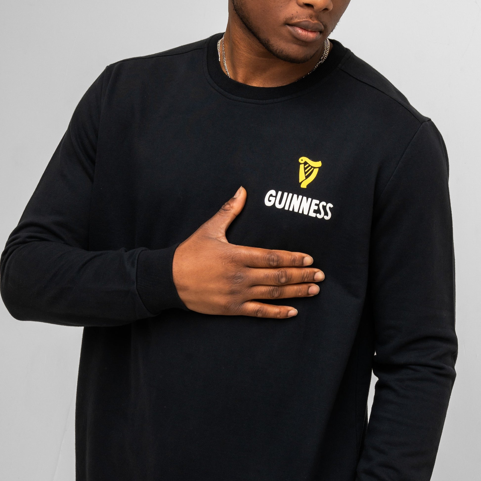 A person wearing the Guinness Seb Curi Lovely Day Sweatshirt in black, featuring a yellow harp logo and "Guinness," rests their hand on their chest. Available from Guinness Webstore US.