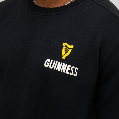Someone wears the Guinness Seb Curi Lovely Day Sweatshirt in black from Guinness Webstore US, featuring a yellow harp logo and "Guinness" print on the chest, inspired by Sebastian Curi's artwork.