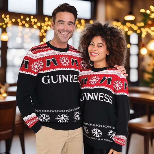 Two people stand indoors, smiling at the camera in matching Official Guinness Holiday Sweaters by Guinness, featuring festive red, white, and black patterns. Cheerful holiday lights twinkle in the background—a perfect seasonal touch.