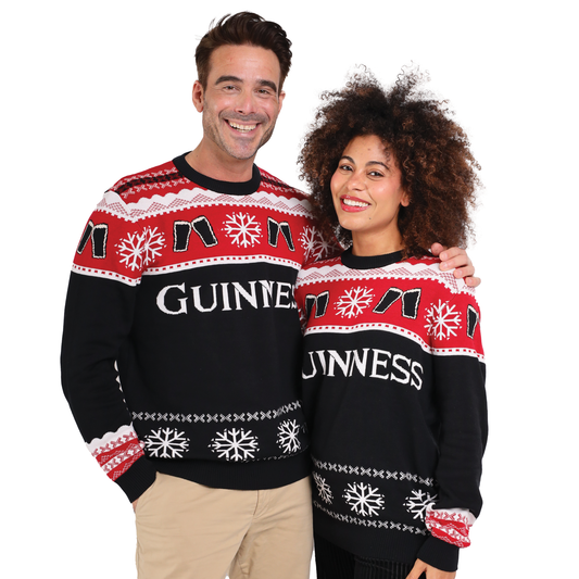 A man and woman wear matching Official Guinness Holiday Sweaters by Guinness, featuring festive black, red, and white snowflake patterns—perfect for bringing cheer to your holiday wardrobe.