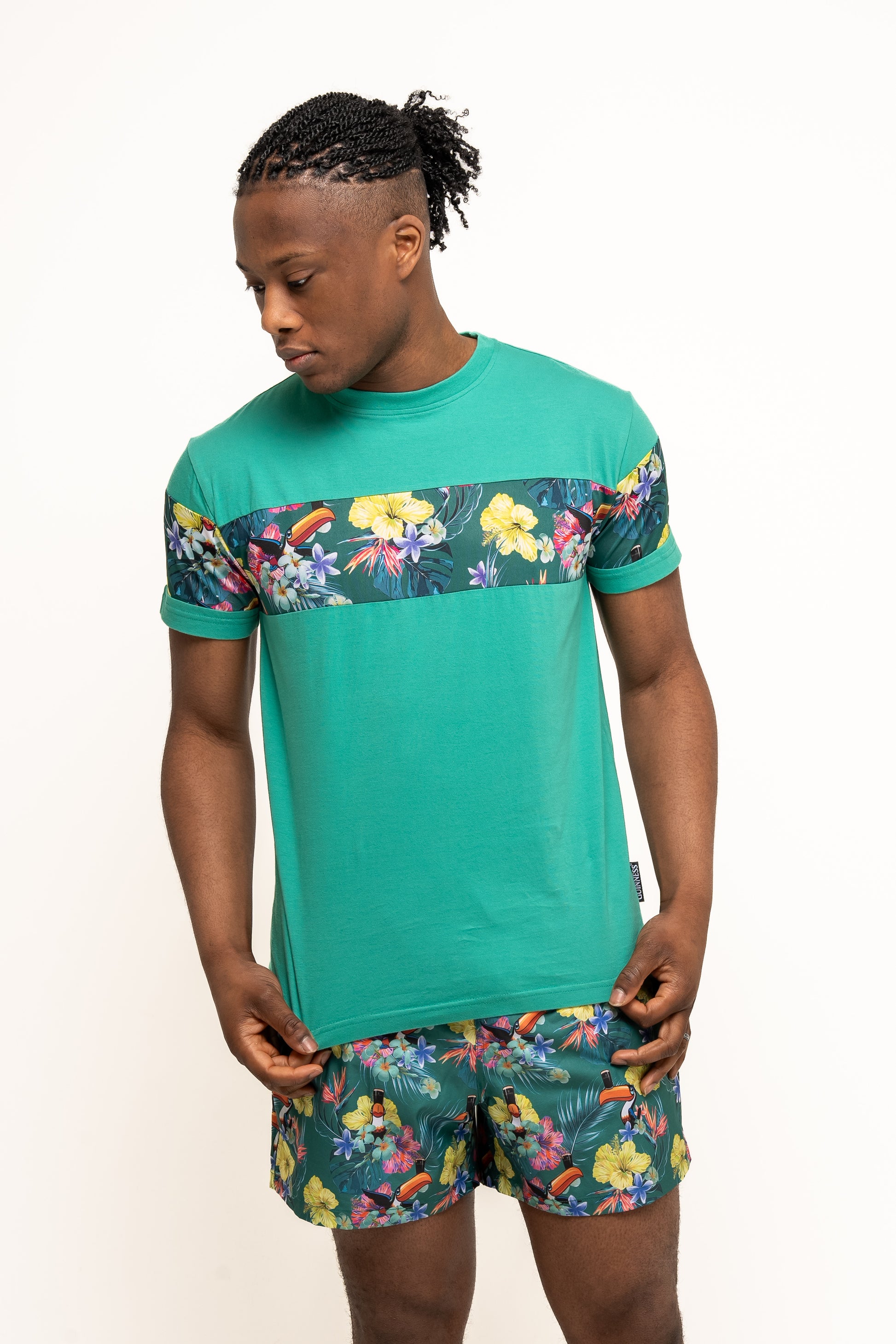 A person stands against a plain white background wearing a teal t-shirt with a floral print stripe and eco-friendly Guinness Toucan Hawaiian Green Beach Shorts from Guinness Webstore US, both featuring matching floral prints.