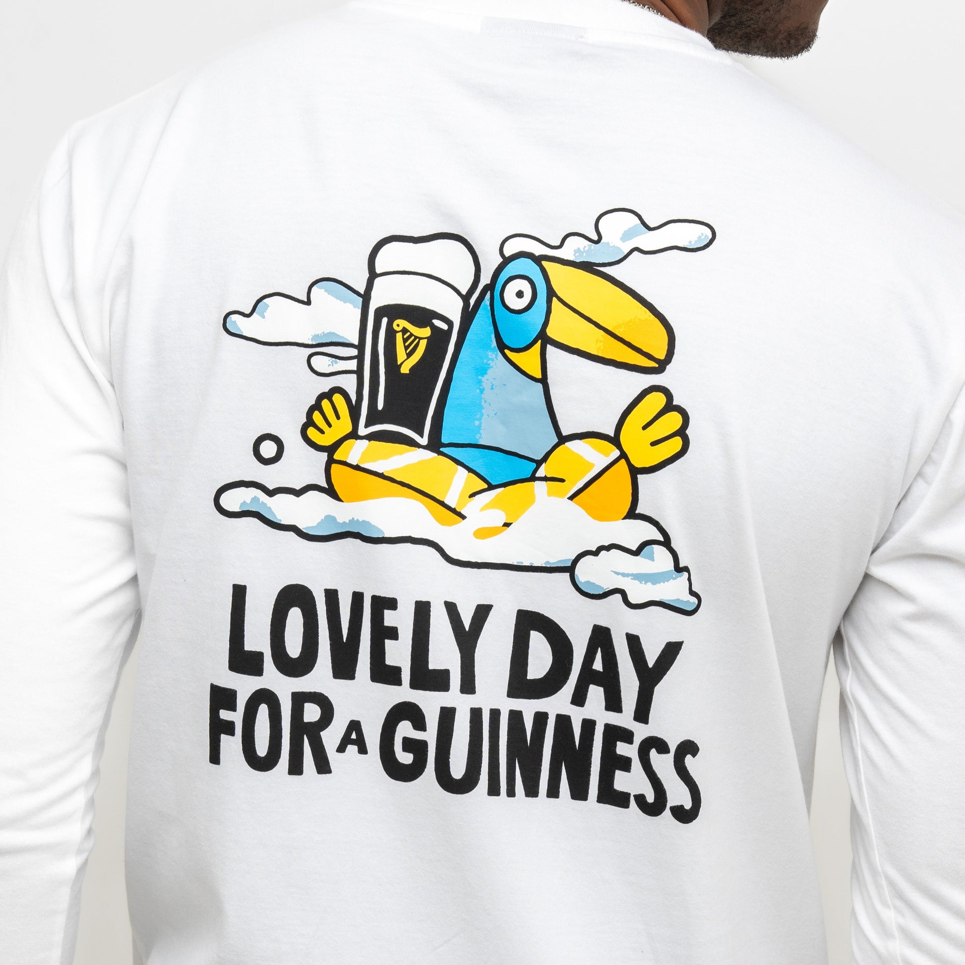 A person wears the Guinness Webstore US Seb Curi Lovely Day Longsleeve Tee in white, featuring a cartoon toucan with a pint and "LOVELY DAY FOR A GUINNESS"—a unique Sebastian Curi collaboration wearable art piece.