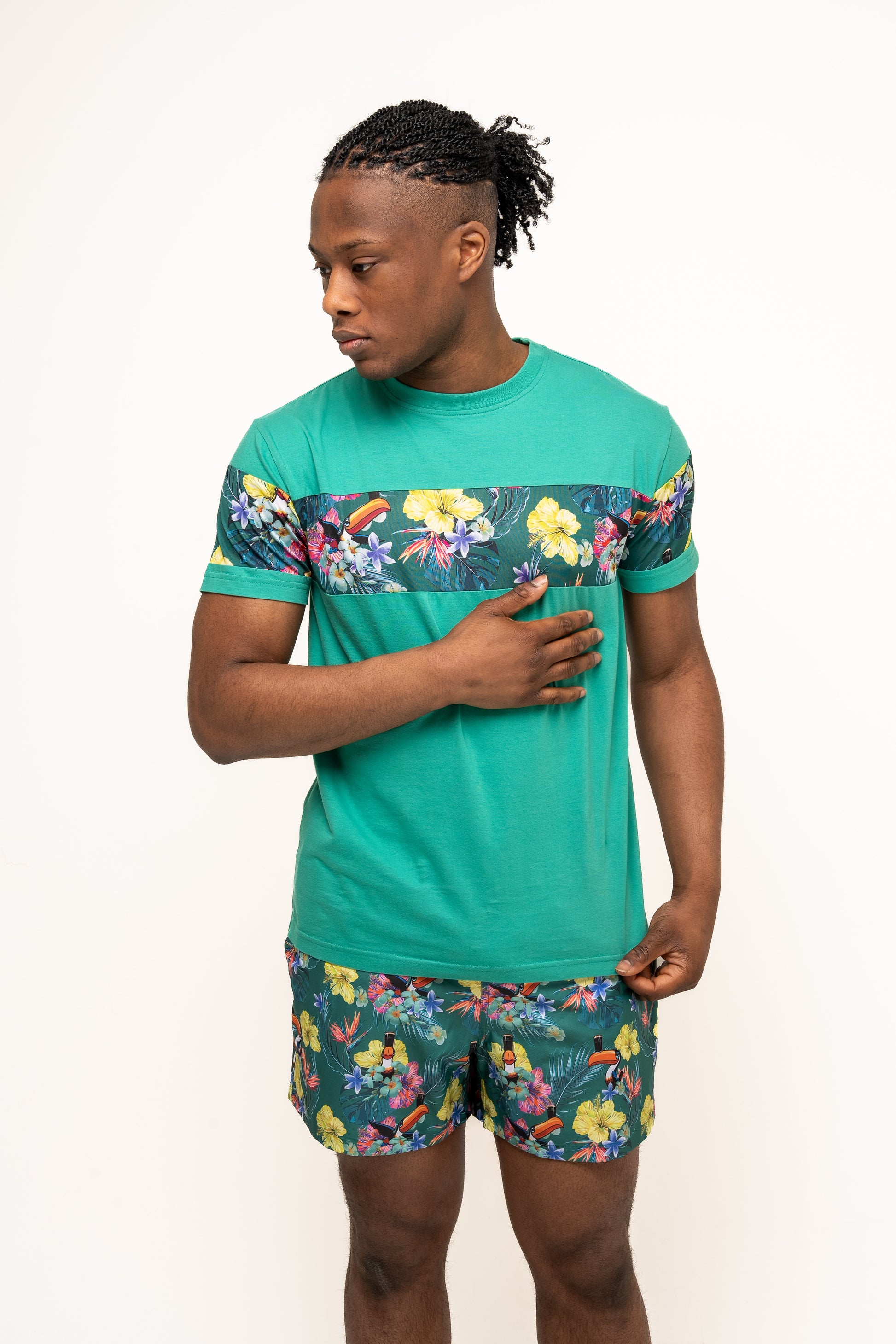 A man stands against a white background wearing Guinness Toucan Hawaiian Green Beach Shorts from Guinness Webstore US, with one hand on his chest and looking to his left.