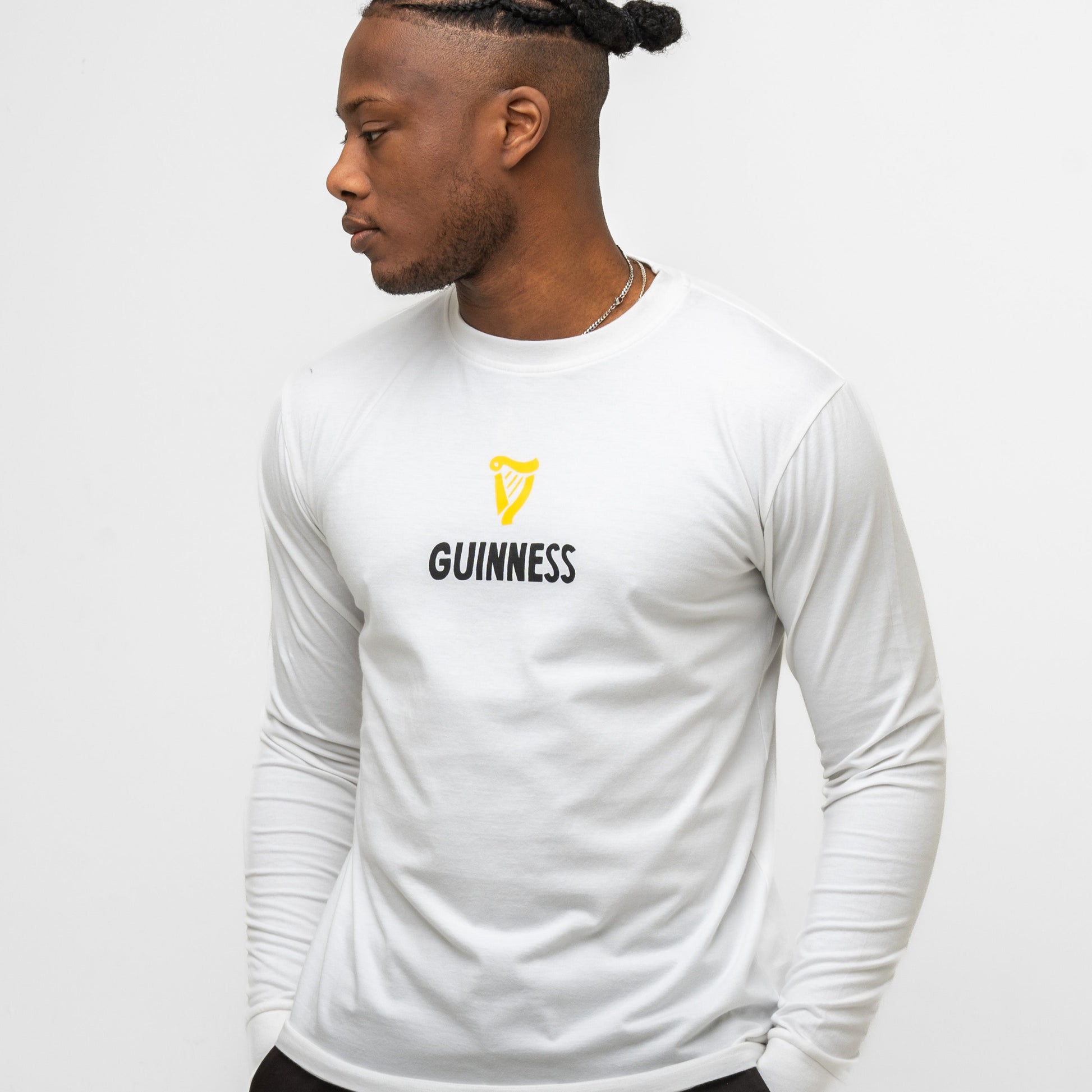 A man wears the Guinness Seb Curi Lovely Day Longsleeve Tee - White from Guinness Webstore US, featuring a yellow harp logo and bold "GUINNESS" text on the front, part of the Sebastian Curi collaboration, against a plain white background.