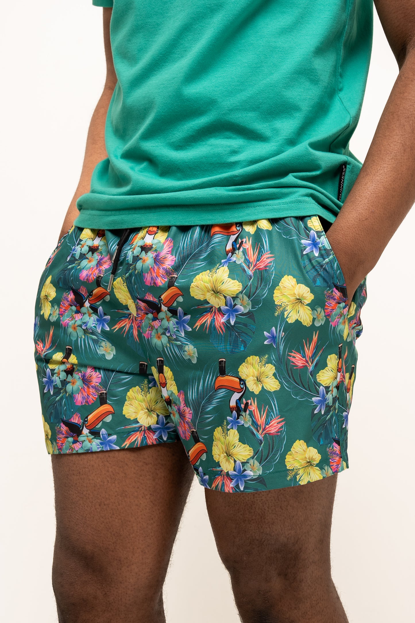 A person wearing a green t-shirt and Guinness Toucan Hawaiian Green Beach Shorts from Guinness Webstore US, hands in pockets, stands against a plain background.