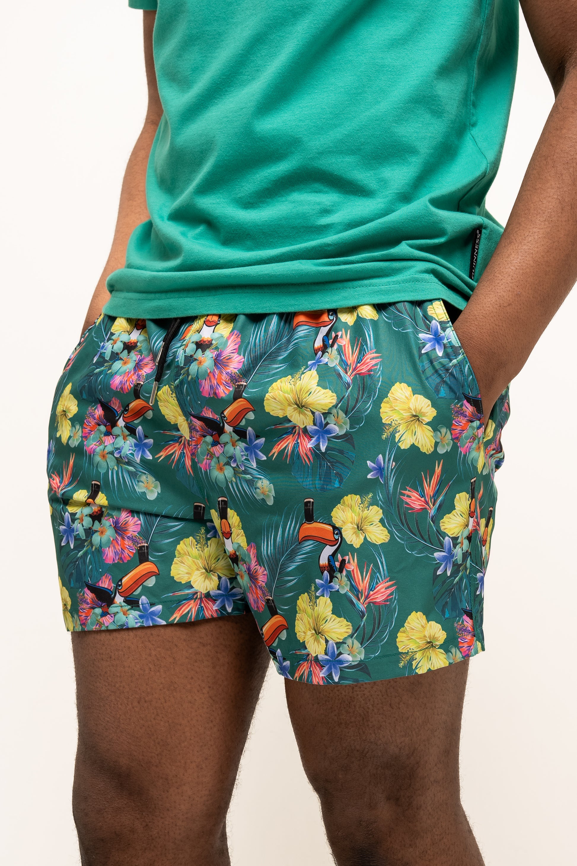 A person wearing a green t-shirt and Guinness Toucan Hawaiian Green Beach Shorts from Guinness Webstore US, hands in pockets, stands against a plain background.