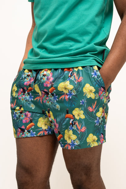 A person wearing a green t-shirt and Guinness Toucan Hawaiian Green Beach Shorts from Guinness Webstore US, hands in pockets, stands against a plain background.