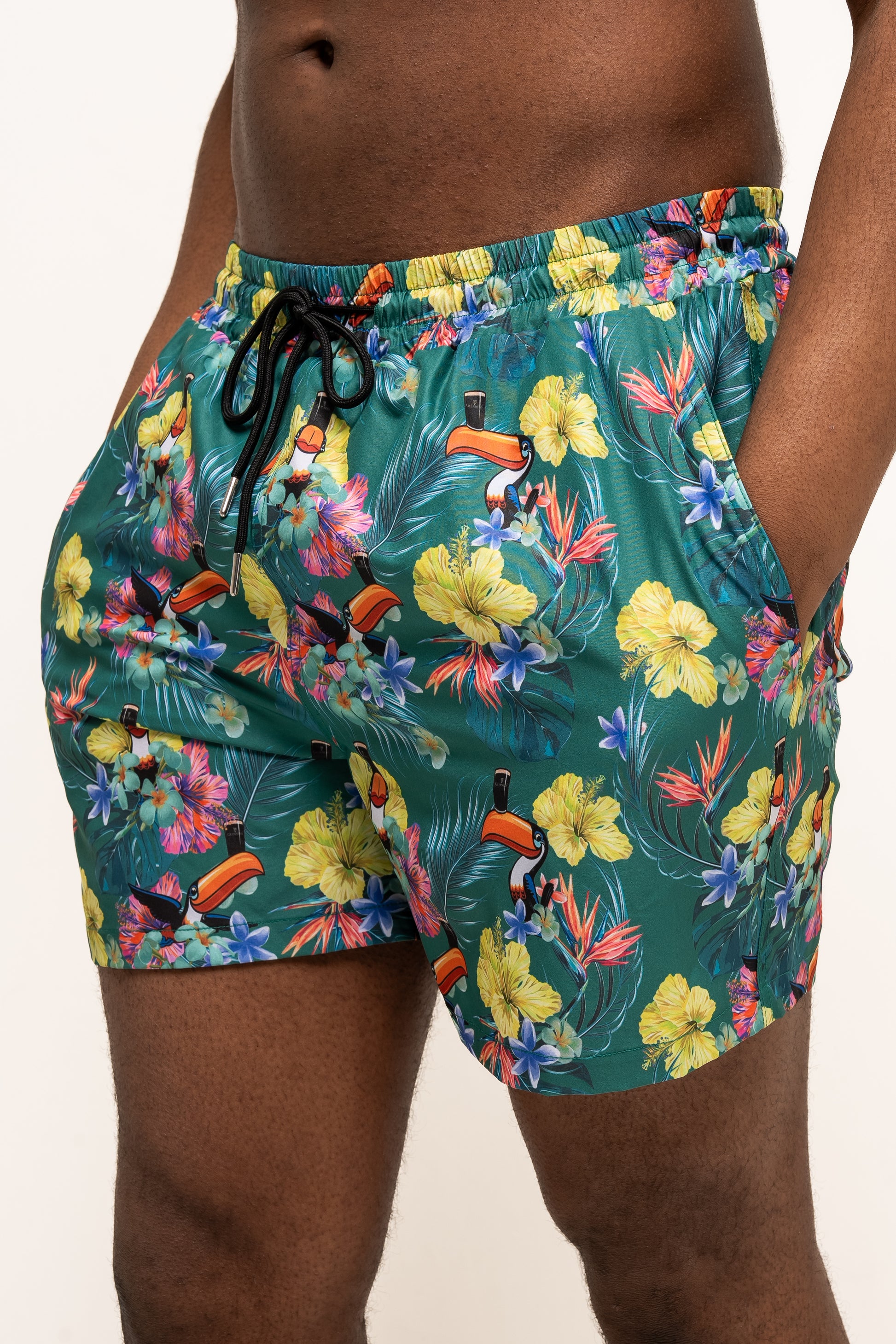A person stands against a plain background wearing Guinness Webstore US’s Guinness Toucan Hawaiian Green Beach Shorts, featuring a vibrant tropical floral and toucan print.