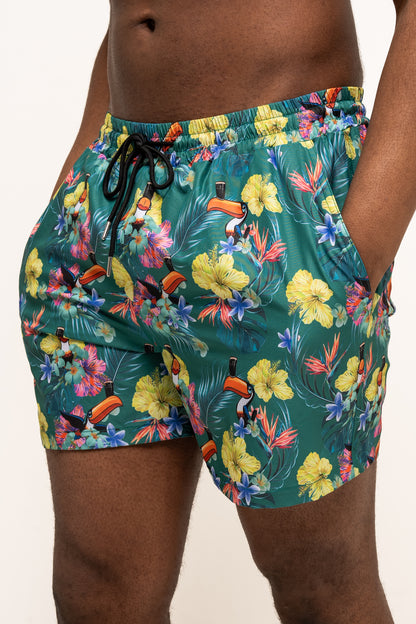 A person stands against a plain background wearing Guinness Webstore US’s Guinness Toucan Hawaiian Green Beach Shorts, featuring a vibrant tropical floral and toucan print.