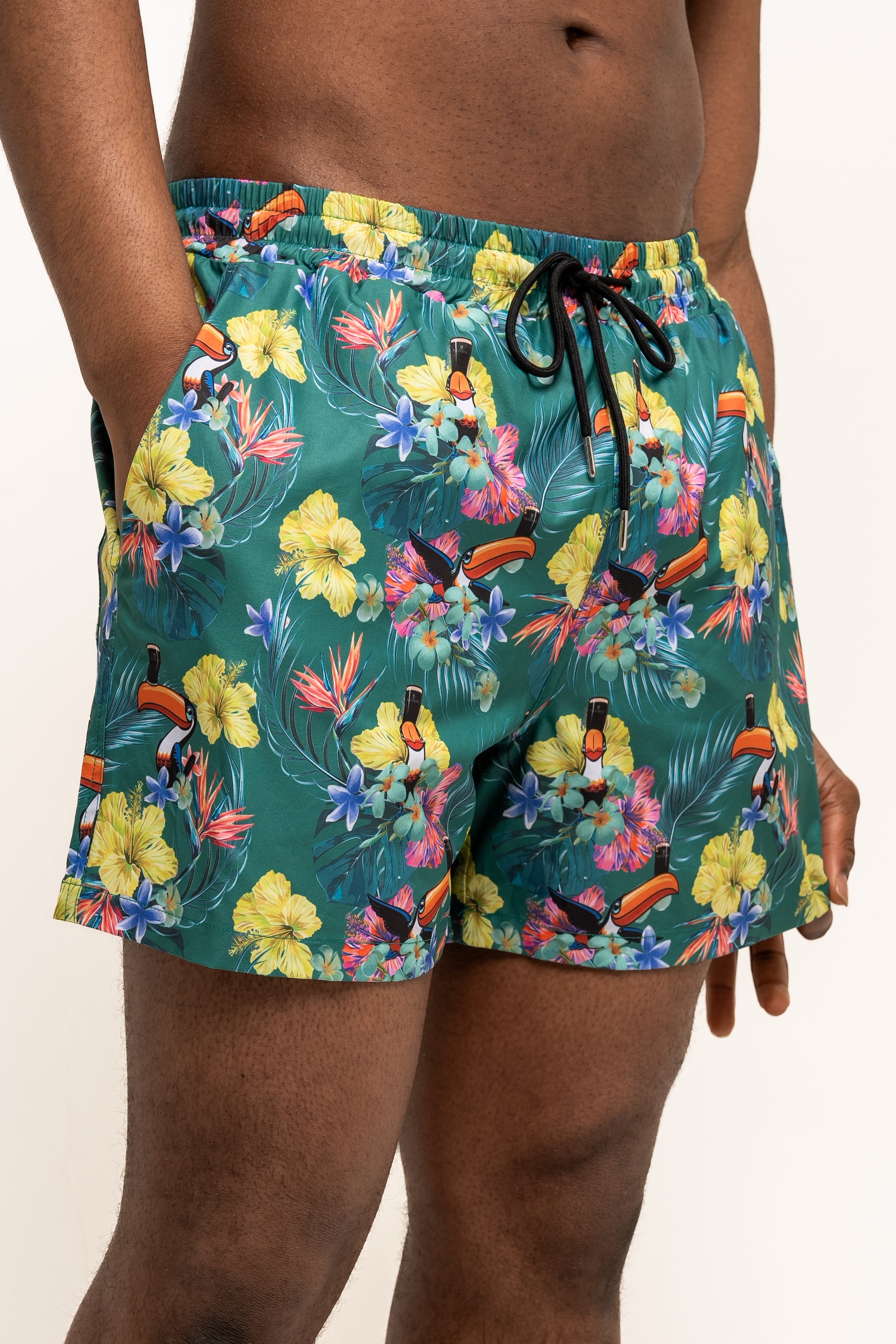 A person wearing Guinness Toucan Hawaiian Green Beach Shorts from Guinness Webstore US, featuring a vibrant tropical floral and toucan print, stands with hands in pockets against a plain background.