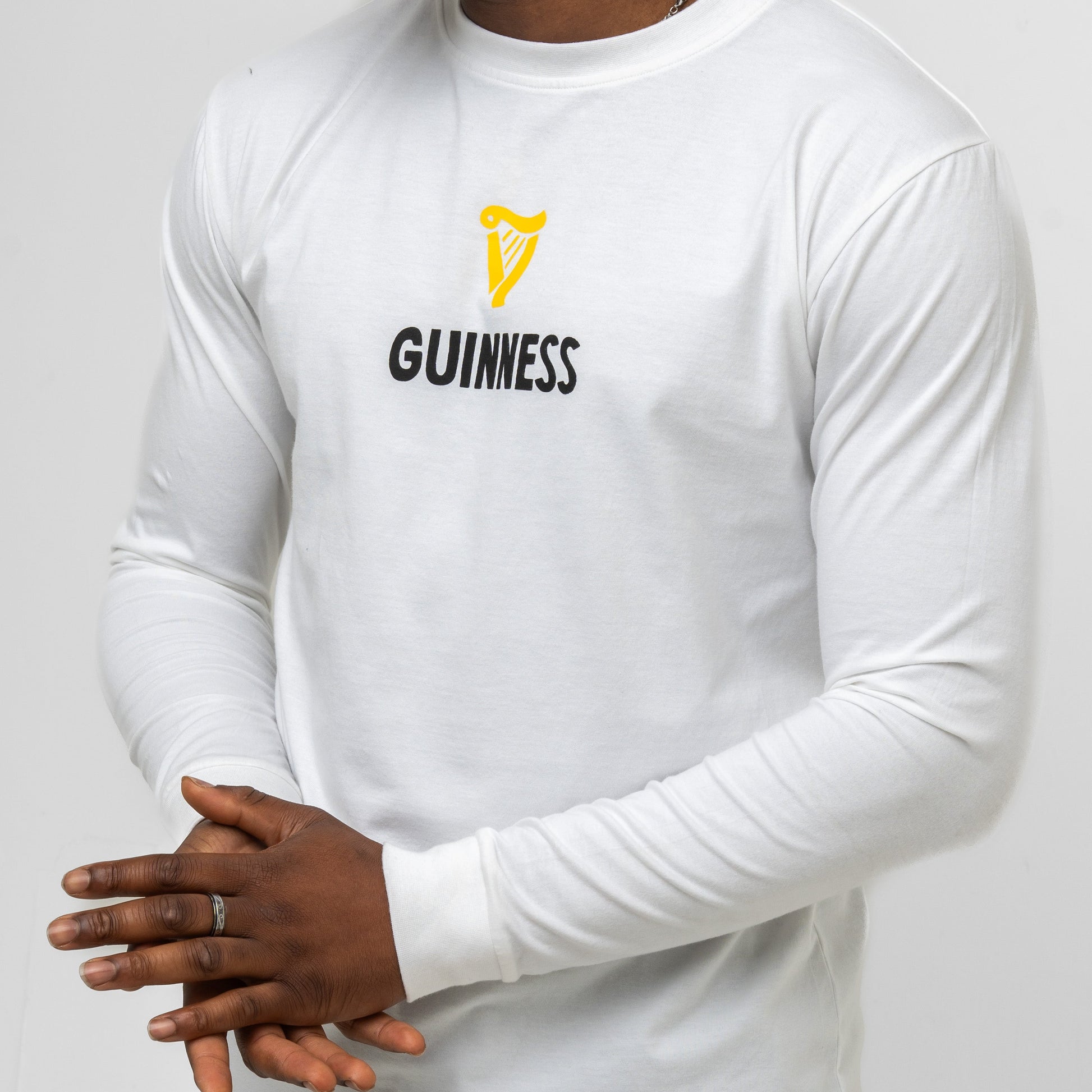 Wearing the Guinness Seb Curi Lovely Day Longsleeve Tee in white from Guinness Webstore US, a person stands against a plain background with hands clasped—highlighting this special collaboration wearable art.