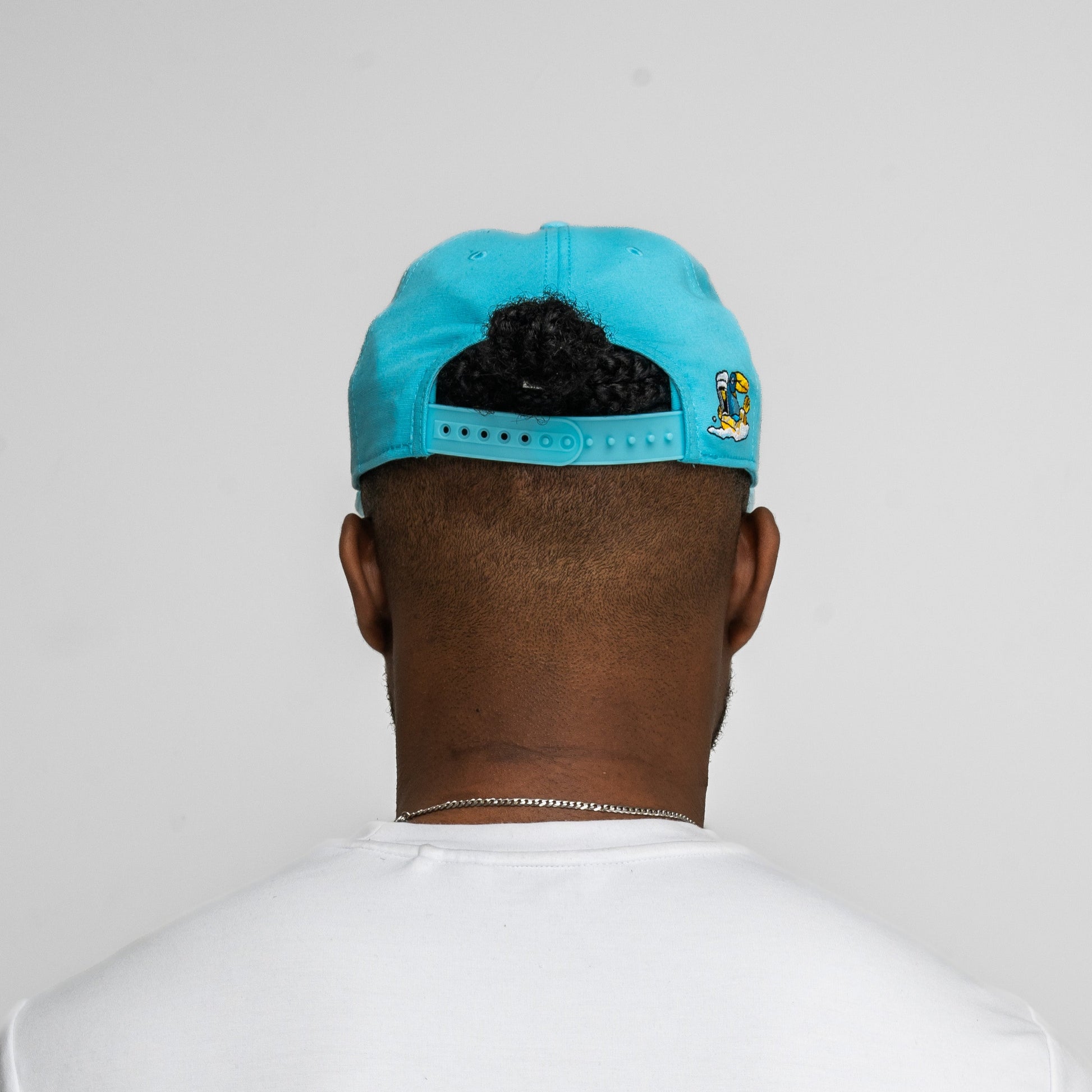 Person wearing a Guinness Seb Curi Lovely Day Flat Brim Cap - Blue by Guinness Webstore US backwards, white t-shirt, and silver necklace, facing away from the camera against a plain gray background.
