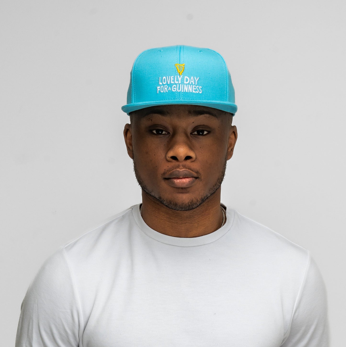 A person wearing the Guinness Seb Curi Lovely Day Flat Brim Cap - Blue from Guinness Webstore US and a white shirt stands against a plain light gray background.