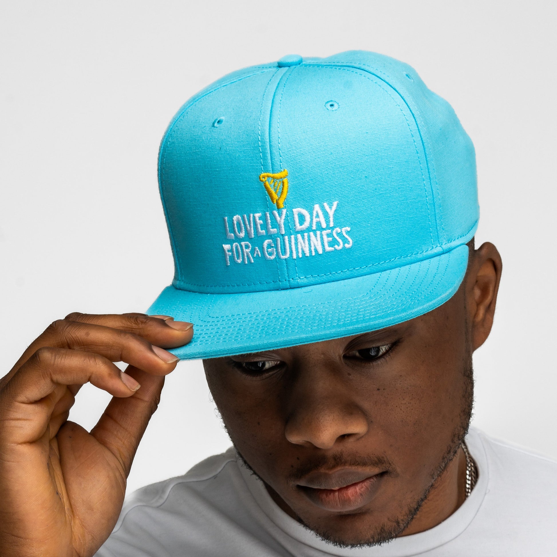 A person adjusts the Guinness Seb Curi Lovely Day Flat Brim Cap in blue from Guinness Webstore US, featuring "LOVELY DAY FOR A GUINNESS" embroidery and a small yellow harp logo on the front.