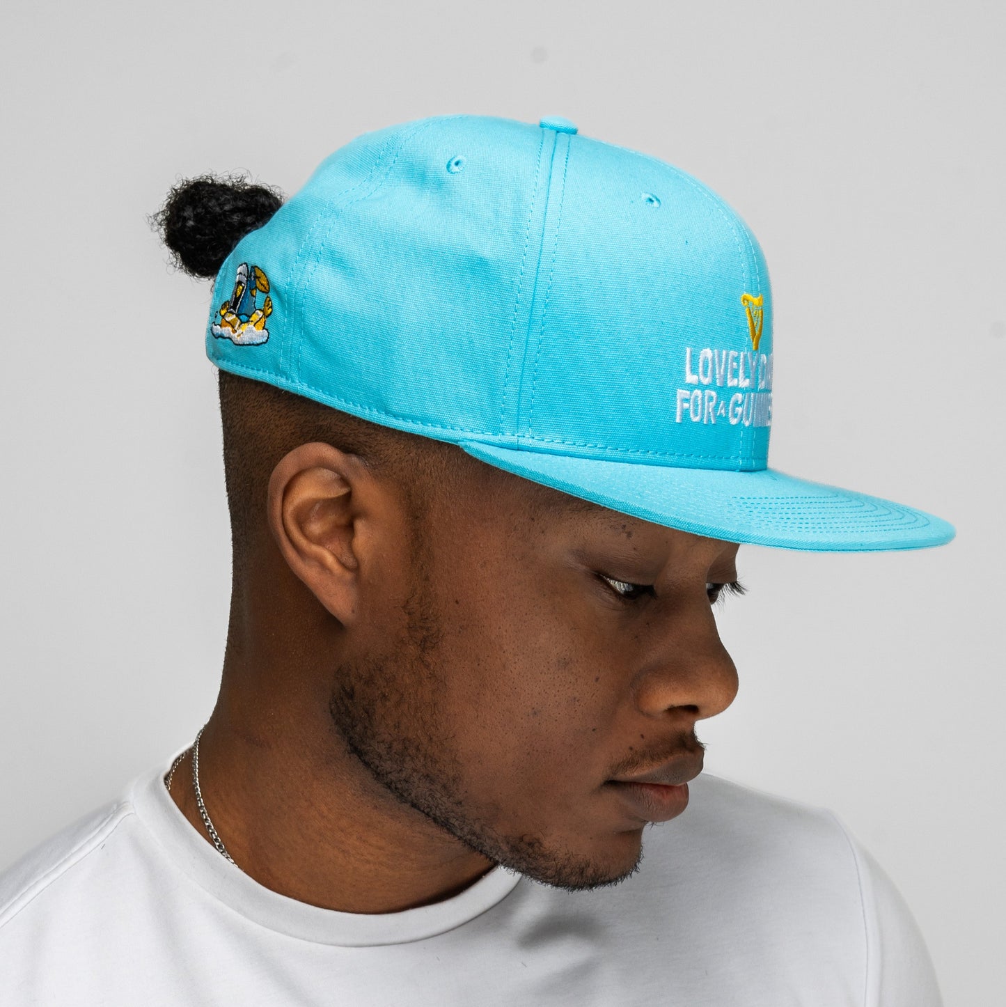 A person wearing a blue Guinness Seb Curi Lovely Day Flat Brim Cap from Guinness Webstore US, featuring embroidered text and a side graphic, facing left against a plain light background.