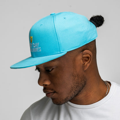 A man wearing a white t-shirt and the Guinness Seb Curi Lovely Day Flat Brim Cap in blue from Guinness Webstore US looks downward against a plain gray background.