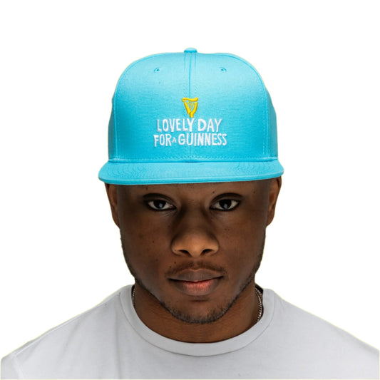 A man faces the camera wearing the Guinness Seb Curi Lovely Day Flat Brim Cap - Blue from Guinness Webstore US, featuring "Lovely Day For A Guinness" embroidered on the front, set against a plain background.
