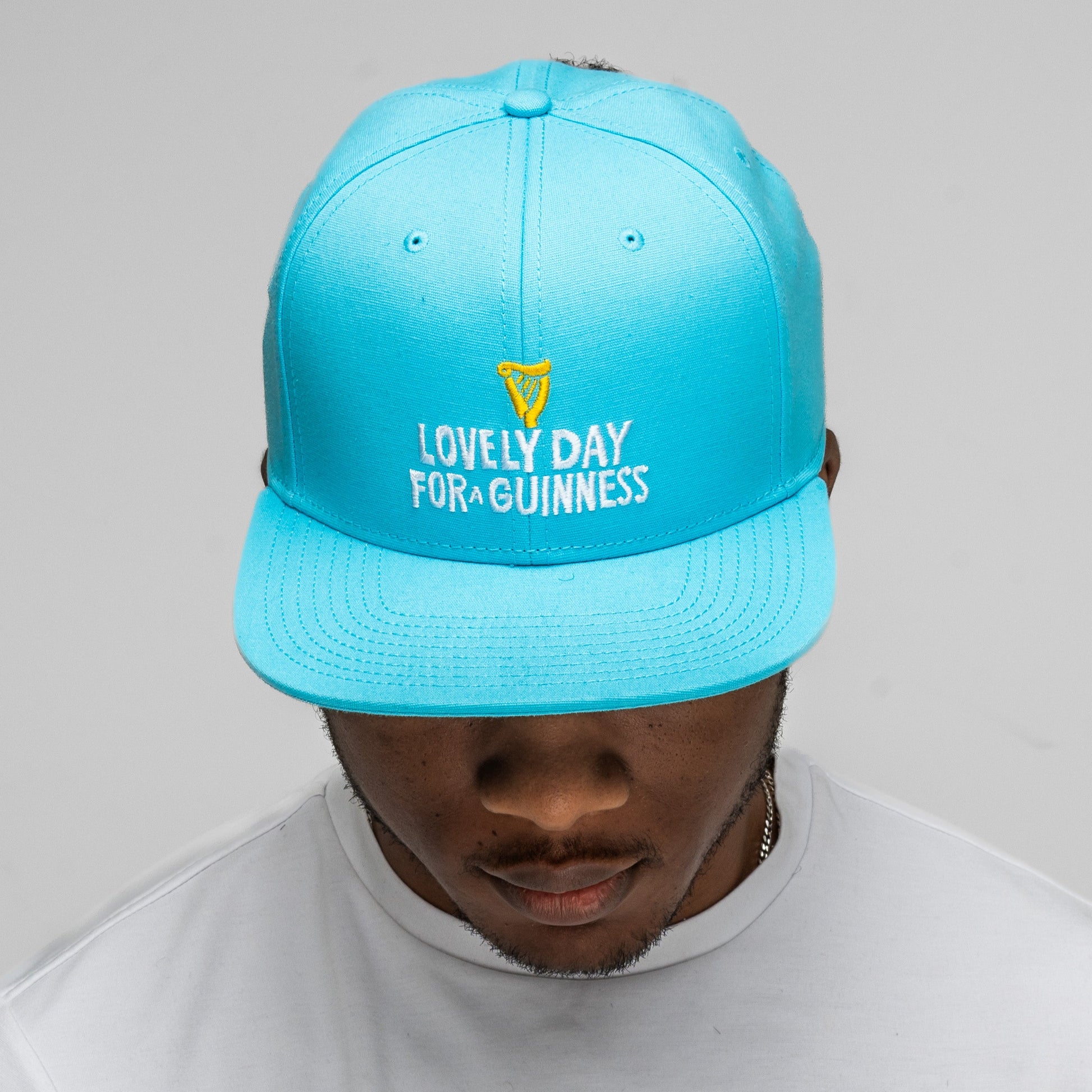 A person wearing the Guinness Seb Curi Lovely Day Flat Brim Cap in blue from Guinness Webstore US and a white t-shirt, looking down against a plain background—echoing Sebastian Curi's playful style.