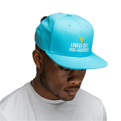 A man wearing the Guinness Seb Curi Lovely Day Flat Brim Cap - Blue from Guinness Webstore US and a white t-shirt looks downward against a plain background.
