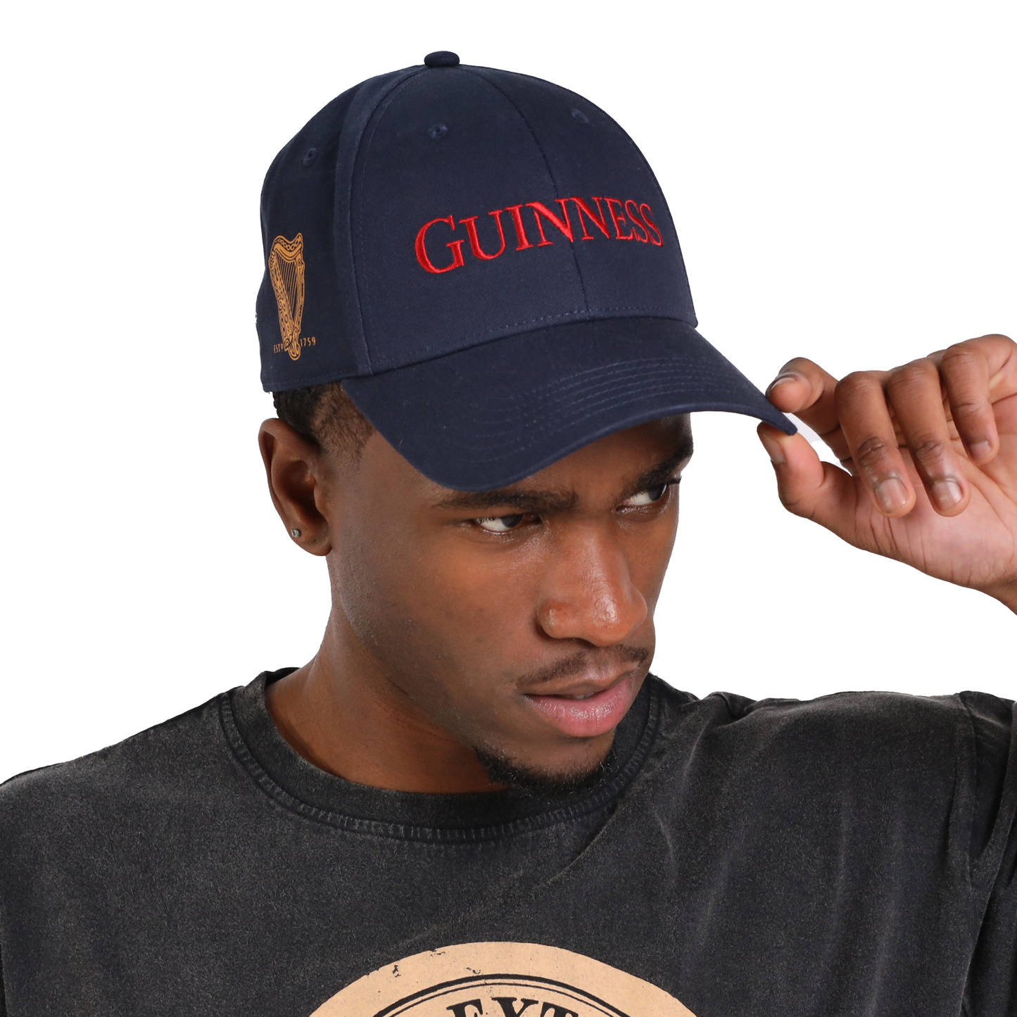 A person wearing a navy blue "Guinness Baseball Cap - Transport Collection" with red lettering, adjusting the brim with their right hand, showcasing its timeless style from the Guinness Webstore US.