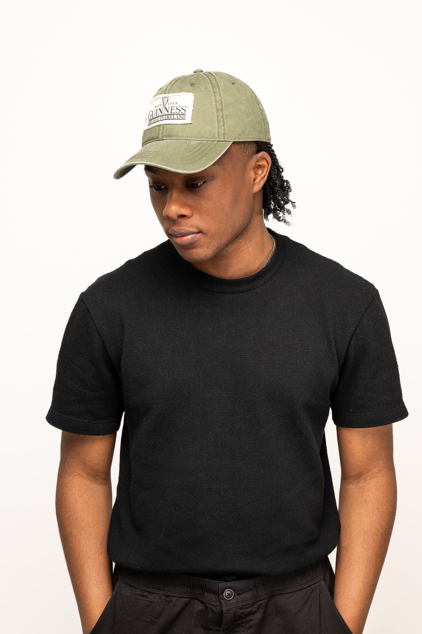 A person in a black short-sleeve shirt and a Guinness Olive Green Patch Cap from Guinness Webstore US stands against a plain white background, looking down with hands in pockets.
