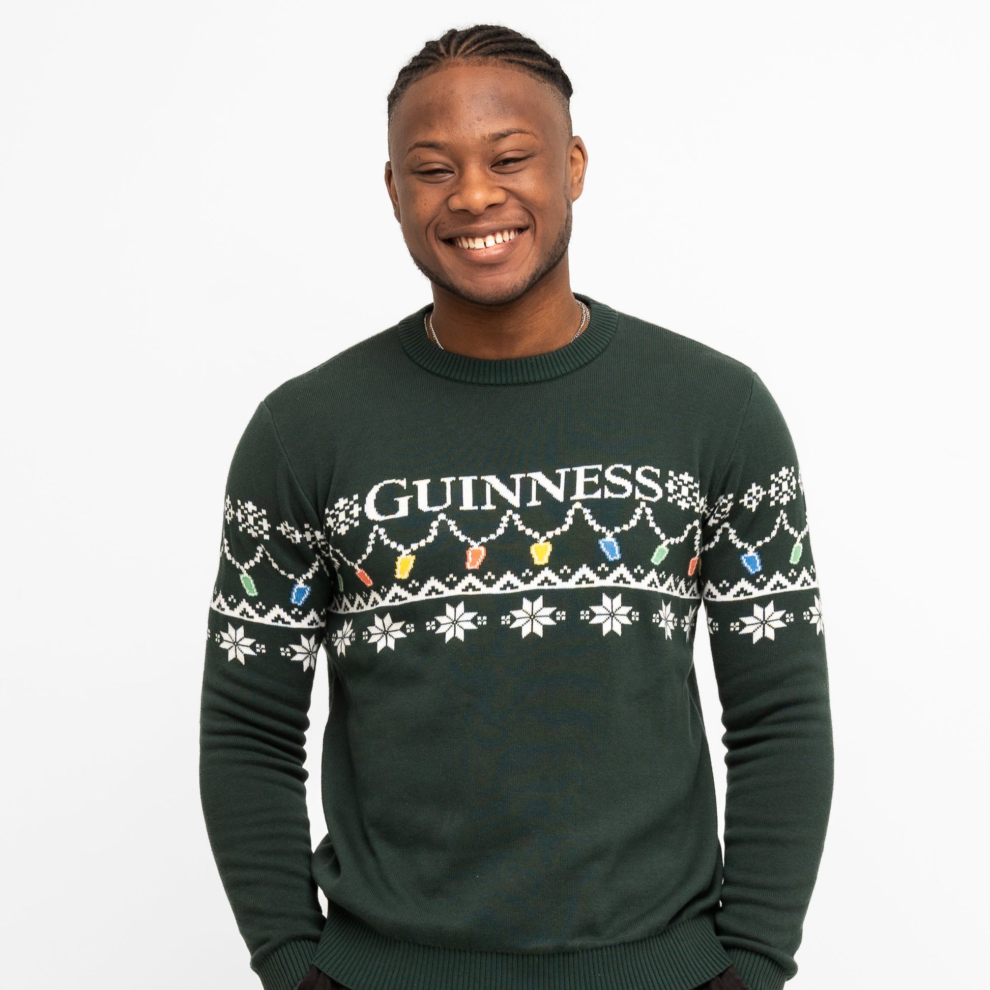 A person smiles wearing the Guinness Holiday Pint Lights Sweater - Green from Guinness Webstore US, featuring "GUINNESS" and festive snowflakes with colored pint lights on a dark green background.