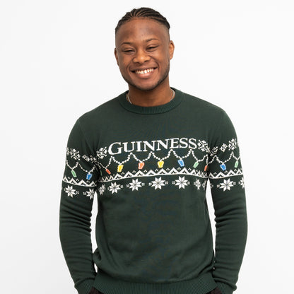 A person smiles wearing the Guinness Holiday Pint Lights Sweater - Green from Guinness Webstore US, featuring "GUINNESS" and festive snowflakes with colored pint lights on a dark green background.