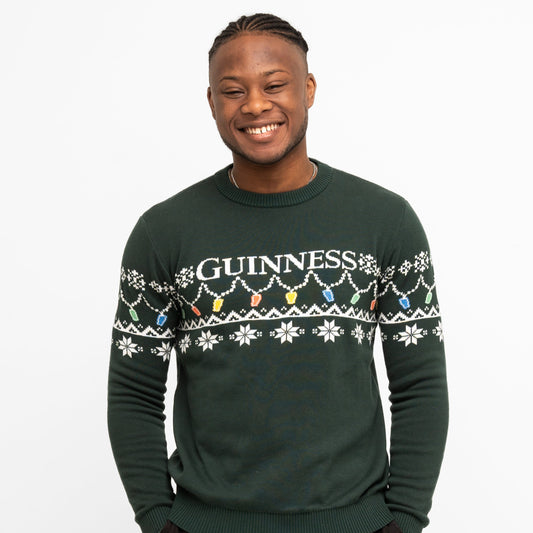 A person smiles wearing the Guinness Holiday Pint Lights Sweater - Green from Guinness Webstore US, featuring "GUINNESS" and festive snowflakes with colored pint lights on a dark green background.
