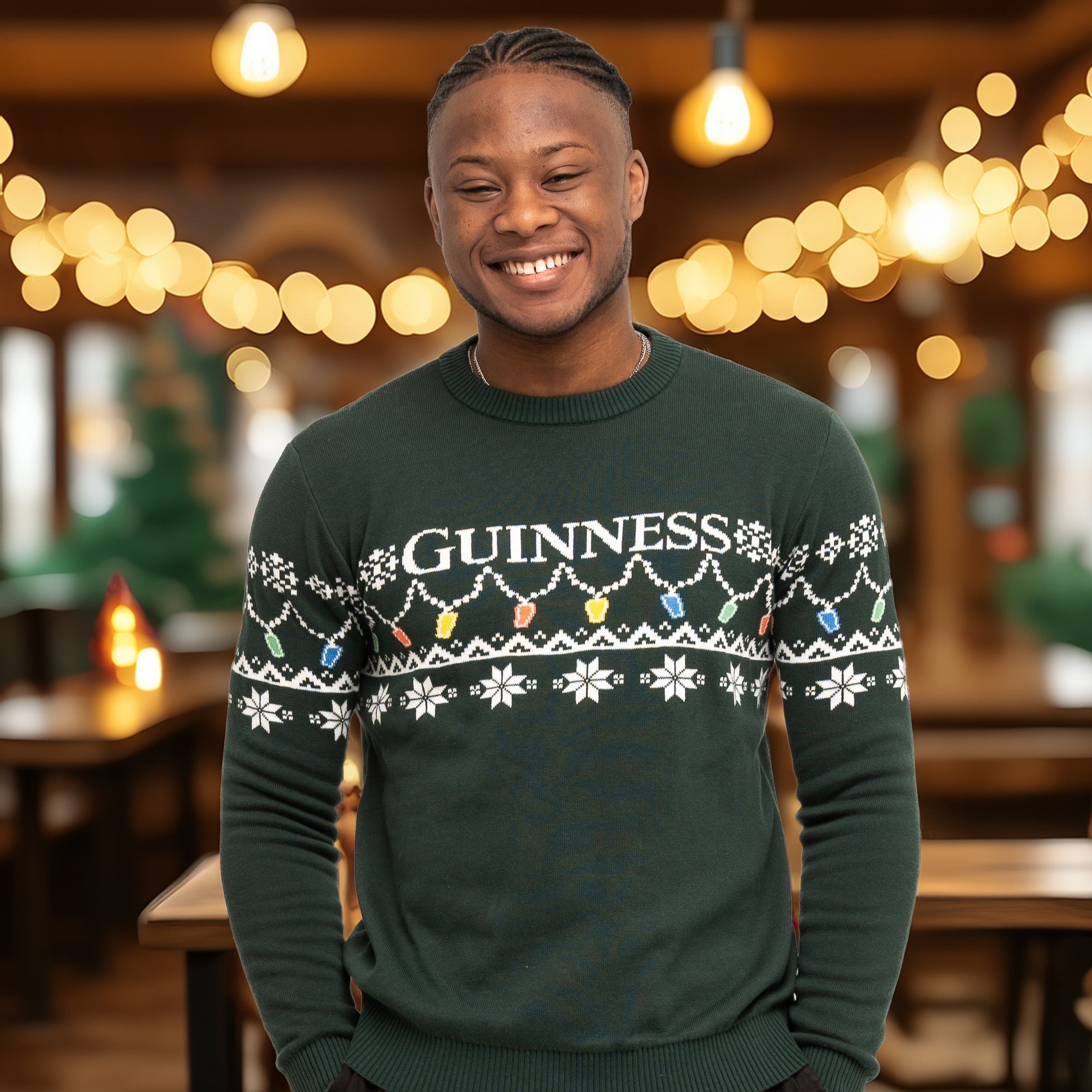 A smiling person wears the Guinness Holiday Pint Lights Sweater - Green from Guinness Webstore US, featuring festive lights and snowflake patterns, while standing indoors with blurred glowing lights in the background.