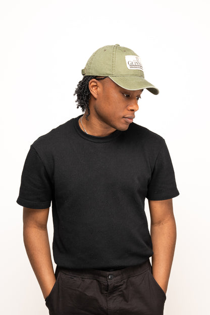 A person wearing the Guinness Olive Green Patch Cap from Guinness Webstore US, along with a black t-shirt and black pants, stands against a plain white background, looking down with hands in their pockets.