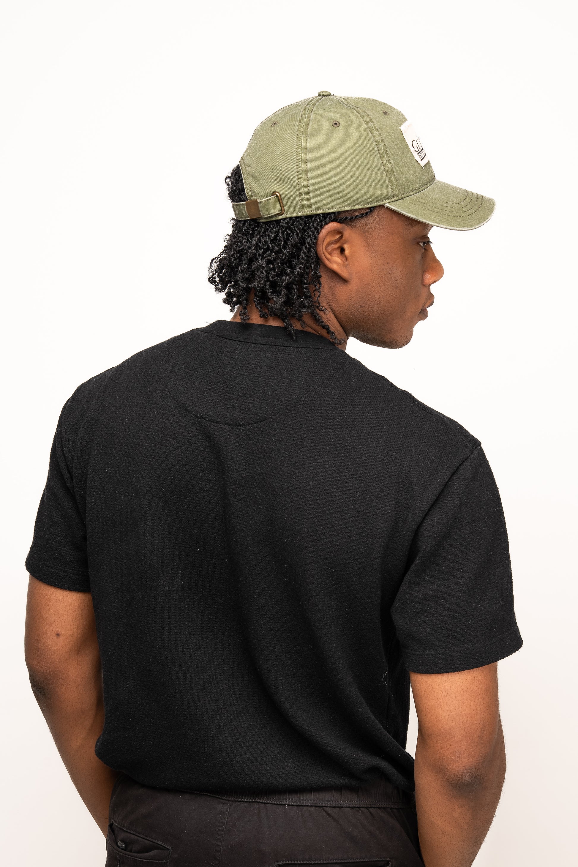 A person with short curly hair wears a Guinness Olive Green Patch Cap from Guinness Webstore US and a black t-shirt, facing away from the camera against a plain white background.