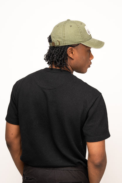A person with short curly hair wears a Guinness Olive Green Patch Cap from Guinness Webstore US and a black t-shirt, facing away from the camera against a plain white background.