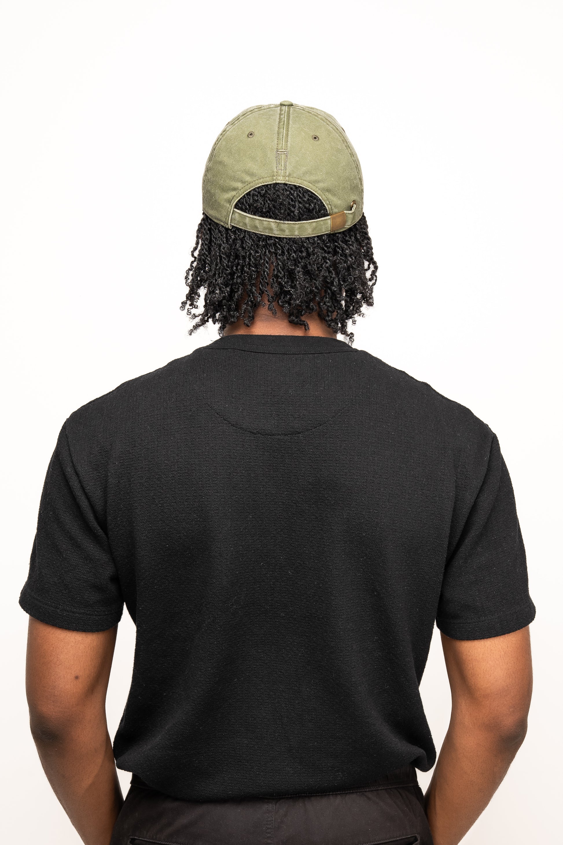 A person with curly hair is shown from behind, wearing a black t-shirt and the Guinness Olive Green Patch Cap from Guinness Webstore US.