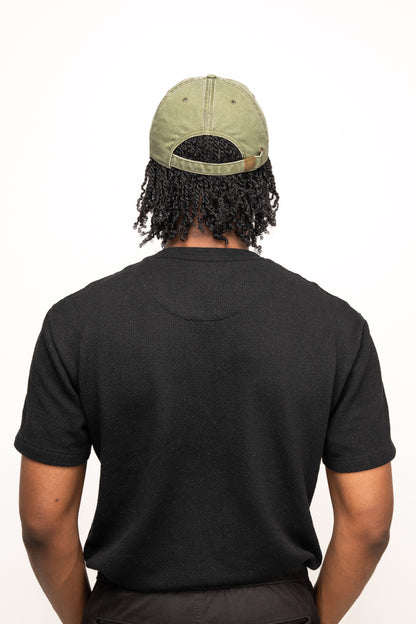 A person with curly hair is shown from behind, wearing a black t-shirt and the Guinness Olive Green Patch Cap from Guinness Webstore US.