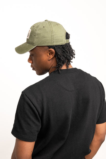 A person wearing a Guinness Olive Green Patch Cap from the Guinness Webstore US and a black t-shirt is shown from behind, looking to the side against a plain light background.