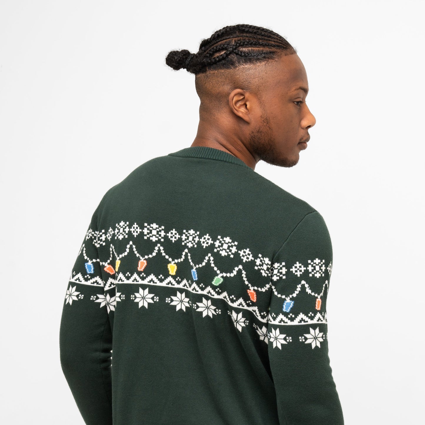 A man with short braids, seen from behind, wears the Guinness Holiday Pint Lights Sweater in green from Guinness Webstore US, featuring festive white snowflakes and colorful holiday light designs.