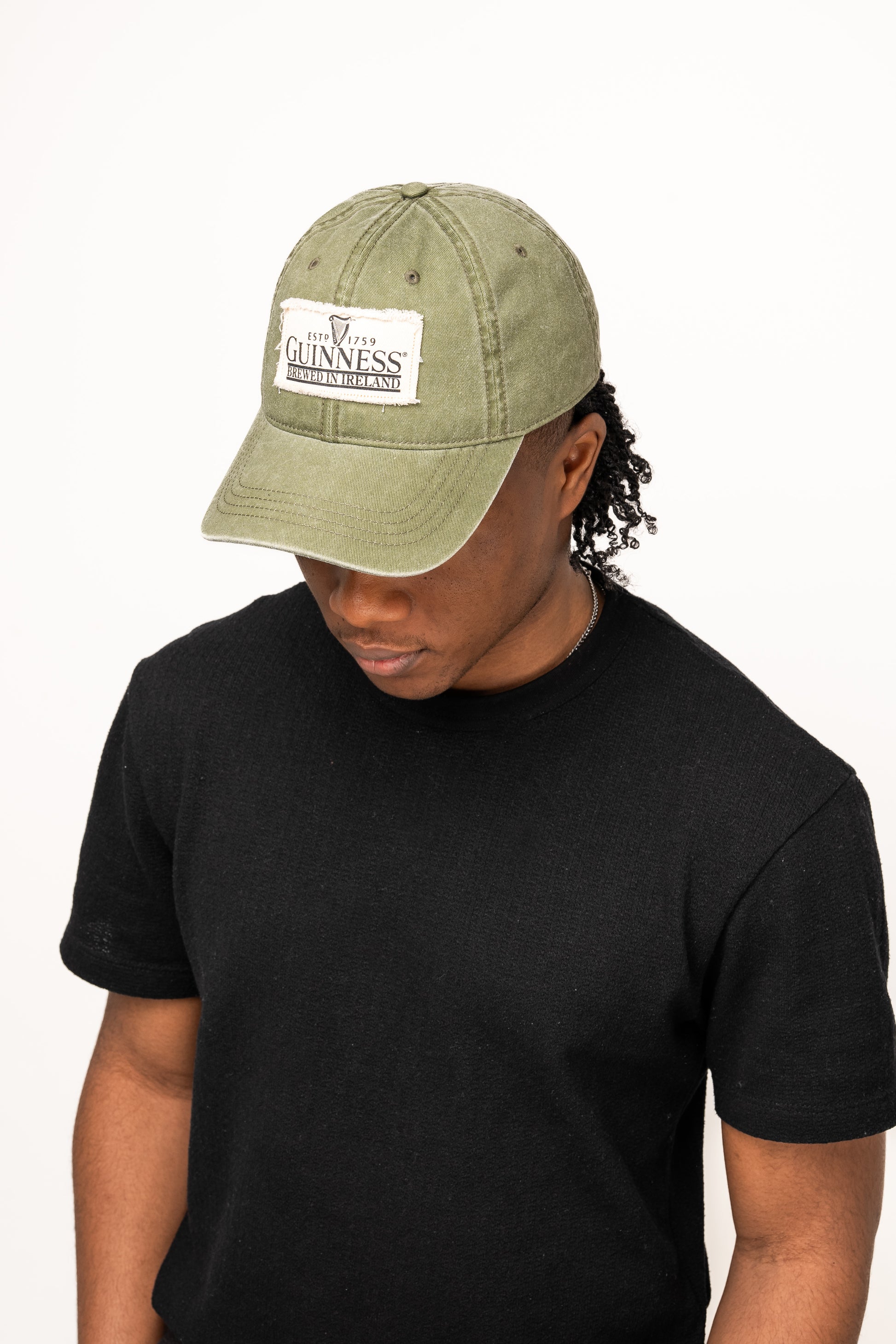 Against a plain white background, a person wearing a black t-shirt and the Guinness Olive Green Patch Cap from Guinness Webstore US looks downward, highlighting classic Irish accessories style.