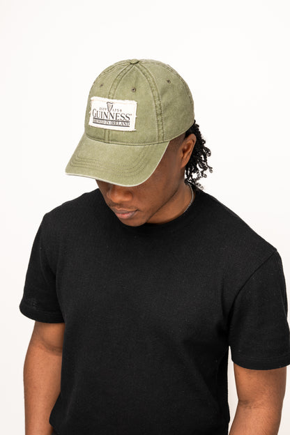 Against a plain white background, a person wearing a black t-shirt and the Guinness Olive Green Patch Cap from Guinness Webstore US looks downward, highlighting classic Irish accessories style.