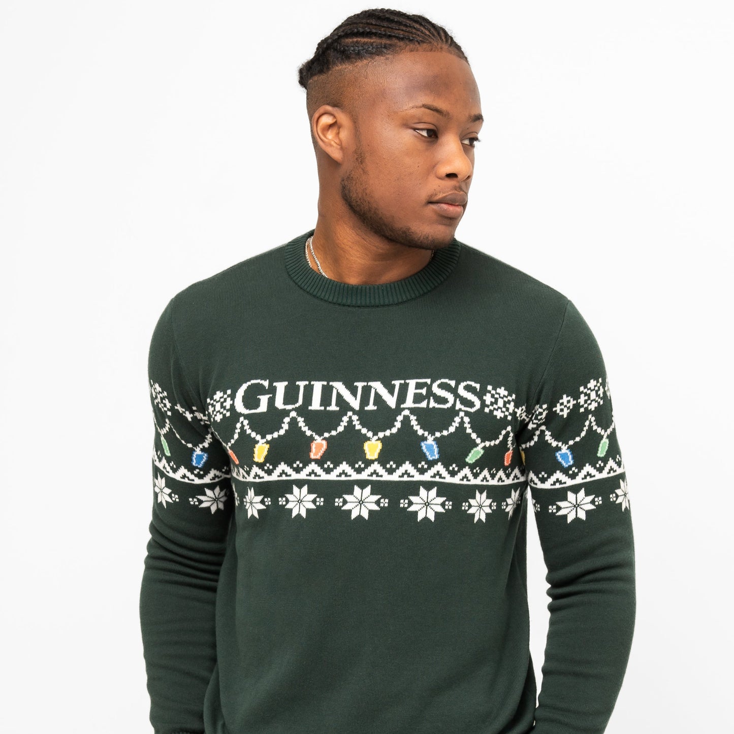 A man wears the Guinness Holiday Pint Lights Sweater - Green from Guinness Webstore US, featuring white snowflakes, colorful lights, and "GUINNESS" across the chest, standing against a plain white background.