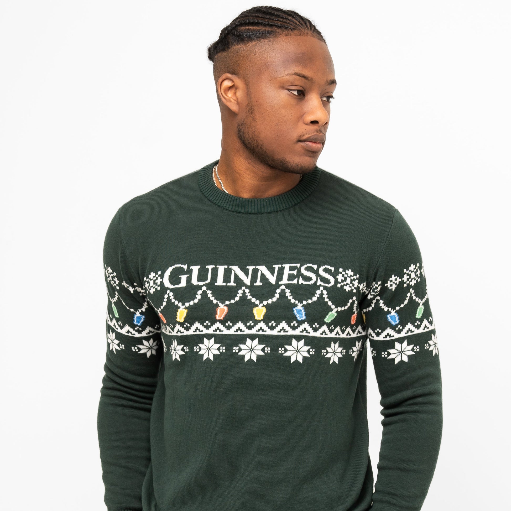 A man wears the Guinness Holiday Pint Lights Sweater - Green from Guinness Webstore US, featuring white snowflakes, colorful lights, and "GUINNESS" across the chest, standing against a plain white background.