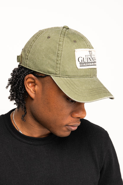 Wearing the Guinness Olive Green Patch Cap from Guinness Webstore US and a black shirt, this person looks down against a plain white background—a timeless tribute to Irish style.